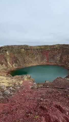 The video shows a high-angle view of a volcanic crater filled with turquoise water. The crater's walls display layers of red, brown, and green vegetation. A small figure of a person can be seen near the water's edge. The foreground is dominated by reddish-brown rocky terrain. The sky is overcast and gray.