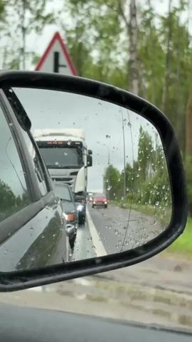 The video shows the side-view mirror of a car stuck in traffic on a rainy day. The mirror is covered with water droplets. Visible in the mirror's reflection are other cars, including a white truck and a red car directly behind the camera's car. The road appears wet and muddy. A road sign with a red triangle and white cross is visible above the mirror. Trees line the road in the background. The sky is overcast. The audio is music.