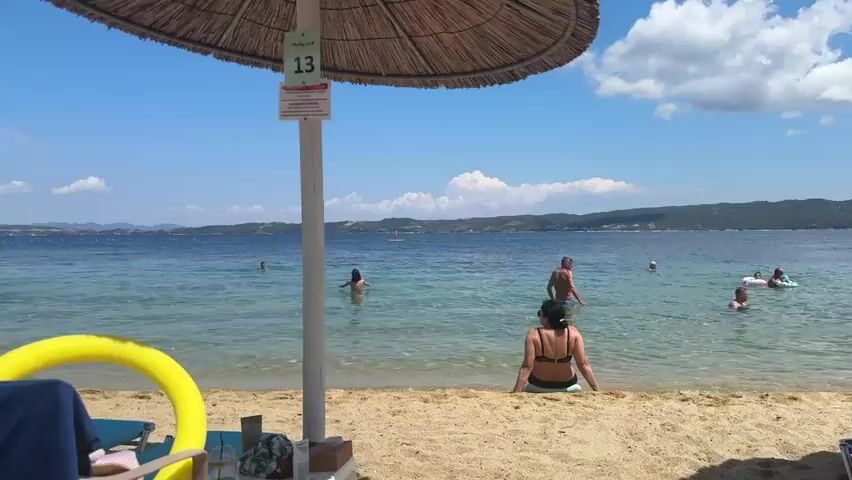 This is a video taken at a beach on a sunny day. The camera is focused on the beach and the sea, with people in the water and on the sand. The video shows a straw umbrella in the foreground, with a sign indicating it is number 13. A woman is sitting on the sand under the umbrella, facing the sea. In the sea, several people are swimming or wading. On the right side of the frame, another umbrella, number 14, is visible, and two people are standing near it, one of whom is wrapped in a blue towel. The sea is calm and the sky is blue with some white clouds. The background shows a coastline with hills. There are also some beach chairs and inflatables on the sand. The video pans slightly to the right, revealing more of the beach and the sea.