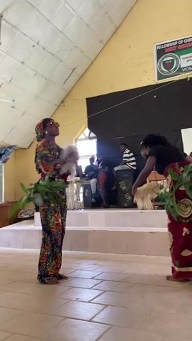 This media file captures a cultural performance in Dutsen Kato, Nigeria. Two young women are the primary subjects, dressed in vibrant, patterned traditional attire. The woman on the left wears a long, flowing dress and headscarf, adorned with colorful floral patterns. She holds a cluster of green leaves in her left hand and a fluffy white object, possibly a fan or ceremonial item, in her right. Her gaze is directed upwards, suggesting engagement with the performance or audience.

The woman on the right is also dressed in traditional clothing, with a dark top and a long, patterned skirt. She too holds a bunch of green leaves and a fluffy white object. Both women appear to be barefoot, standing on a tiled floor. They are positioned on what seems to be a raised platform or stage, indicated by the tiered steps behind them.

In the background, several individuals are seated, possibly musicians or an audience, with instruments visible. To the right, a large black speaker with white lettering that appears to read "FEN DIVA" is noticeable. Behind the performers and audience, a black backdrop is set up. Above this, a yellow wall is adorned with a circular logo and text that reads "FELLOWSHIP OF C..." and "NEXT GENERATION." The ceiling is made of white corrugated material, and natural light streams in from a window on the left. The overall atmosphere seems to be one of cultural celebration and performance, possibly in a community hall or church. The lighting suggests it is daytime.