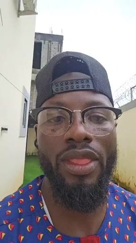 A medium close-up shot shows a Black man wearing a black baseball cap backward, glasses, and a blue shirt with a red and yellow pattern. He has a beard and is looking directly at the camera. The background is blurred but appears to be an outdoor setting with a white wall on the left and a wire fence in the distance. The lighting is overcast.