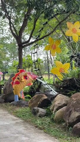 The video shows an outdoor scene with a decorative water feature. A large, colorful, golden koi fish sculpture is positioned to the left, appearing to "spit" water into a small, rocky stream. Behind the fish and stream, there is a large, wheel-like structure decorated with yellow, petal-shaped ornaments that resemble flowers. The wheel is supported by thin black struts. The environment is lush and green, with trees and foliage visible in the background. Large rocks are scattered around the base of the water feature. The ground in the foreground is a paved pathway.