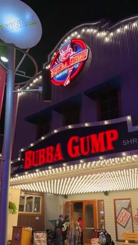 The video shows the exterior of a Bubba Gump Shrimp Co. restaurant at night, located at Citywalk Orlando. The building has a corrugated metal facade, lit by rows of string lights. A large, neon sign featuring a red shrimp figure and the restaurant's name is prominently displayed above the entrance. A few people are visible entering the restaurant, including an individual with long dreadlocks. A sign saying 'Take my Forrest, I'm going home' can be seen on the right.