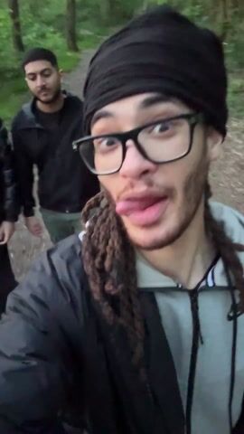 The video captures a selfie perspective of a young man with dreadlocks, glasses, and a black beanie, who is sticking out his tongue and making exaggerated facial expressions. He is wearing a black and gray jacket. Two other men are walking slightly behind him on a dirt path through a wooded area. The path appears to be a hiking trail. The background is filled with green foliage from the trees. The video is slightly shaky, suggesting it was taken while walking.