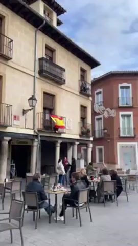The camera pans slowly across a European town square on an overcast day. Several people sit at tables outside a restaurant under a building with a Spanish flag hanging from a balcony. The camera then pans to show other buildings around the square, ending on a large, white church with a tall steeple, a sculpture, and a police car parked nearby.