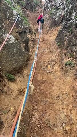 The video shows a person climbing down a steep, rocky slope using ropes for assistance. The slope is reddish-brown in color with some greenery scattered throughout. At the top of the video, a person can be seen further up the slope, dressed in dark shorts and a red shirt with a backpack. Three ropes are strung down the slope, a pink rope, a blue and white rope, and a cream rope. The camera angle changes to a lower perspective, focusing on the ropes and the rock face. A hand covered with a blue glove appears briefly, grabbing one of the ropes. The camera then pans to show the view from the slope, revealing a beach below with red sand, the ocean, and a rocky outcropping in the water.