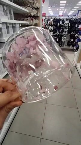 A hand holds a clear glass mug filled with pink flower petals. The mug is being rotated, showing the petals swirling inside. The background appears to be a retail store with shelves stocked with various items, including mugs and clothing. The lighting is bright, typical of a commercial setting.