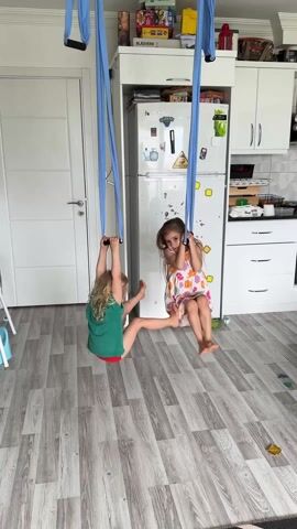 Two young girls are playing in what appears to be a kitchen or an open-plan living space. Both girls are engaged with blue aerial silks or hammocks that are suspended from the ceiling.

At the beginning of the video, the girl on the left, with blonde hair, a green t-shirt, and red shorts, is sitting in her blue silk swing, holding onto the fabric above her head. She appears to be gently swinging. The girl on the right, with long dark hair and a light-colored floral dress, is also sitting in her blue silk swing, holding onto the fabric, and looking directly at the camera with a slight smile.

As the video progresses, the girl on the left pushes off the floor with her feet, lifting herself slightly. She then lets go of the silks and falls to the wooden floor, landing on her bottom with a soft thud, immediately looking up at the camera with a big smile.

Simultaneously, the girl on the right, still in her swing, lifts one leg high into the air, showcasing her flexibility. She then quickly brings her leg down, pushes off the floor with her feet, and stands up, still holding onto the silks. She smiles at the camera as she stands.

The background shows a brightly lit room with a white door on the left and white kitchen cabinets and a large white refrigerator on the right. There are several magnets and stickers on the refrigerator door, including a yellow triangular sticker and a red circular sticker, though the text on them is not clearly legible. A box labeled "BLASVERK" is visible on top of the refrigerator. The floor is covered with light-colored wooden laminate planks. The overall atmosphere is playful and joyful, as the girls seem to be having fun with their aerial swings.