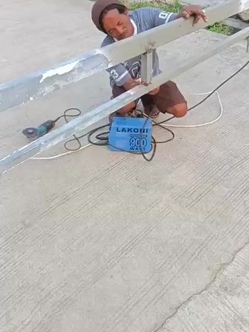A man is kneeling on a concrete surface and welding a metal railing. He is wearing a brown hat, a gray t-shirt, and brown shorts. A welding machine, a blue box with "LAKONI 900 WATT" written on it, is on the ground near him. An angle grinder is also on the ground to his left. The railing appears to be made of metal pipes and has some white paint on it. The background is out of focus but appears to be a grassy area.