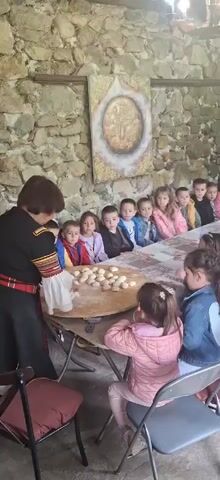 A woman in traditional Bulgarian garb is preparing dough at a large round table surrounded by children. The woman stands on the left side of the table. She is dark-haired, has short bangs, and wears a black and red dress with embroidered sleeves and a red sash. On the table are several dozen small, pale dough balls. A group of children sit at the table, arranged in a row behind the woman. The children are of various ages and wear casual clothing. Two younger girls are sitting off to the right side of the table, facing away from the camera. The environment appears to be inside an old stone building. There is a painting of a golden circle with a cross in the center on the stone wall behind the children.