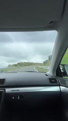 A point-of-view shot from inside a car shows a winding road through a green, hilly landscape under an overcast sky. The dashboard of the car, possibly an Audi given the "AUDI" text, is visible in the foreground, with the "AIRBAG" label on the passenger side. A Coca-Cola cup is seen on the left side of the dashboard. The car is moving, indicating a drive through the countryside.