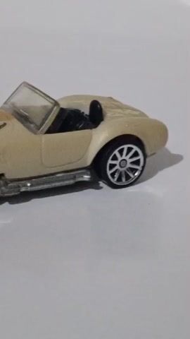 A close-up, eye-level shot focuses on the right side of a toy car. The car is a cream-colored convertible with flame decals on the hood and fender. It has black interior seats, a clear windshield, and silver rims with black tires. The car sits on a white surface, casting a shadow on its right side. The camera slowly pans from the rear of the car to the front.