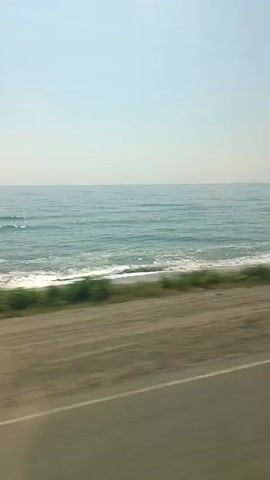A blurred video shows a view from a moving vehicle. The scene consists of a calm, blue ocean on the left, a sandy beach with some green vegetation in the middle, and a road with a white line on the right. The sky is clear and light blue. The motion blur suggests the video was taken from a car or bus traveling alongside the coast.