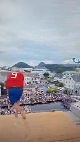 An aerial shot shows a person in red and blue diving into water from a platform. A crowd of people are gathered on the platform and along the waterfront watching. Boats are docked near the platform, and buildings line the shore in the background. The water has a splash from the dive.