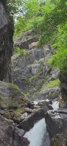 The video captures a dynamic shot of a waterfall cascading down a rocky gorge. No people or visible text are present.

The scene is dominated by a powerful stream of white, frothy water, which tumbles over a series of dark grey and reddish-brown rocks, forming a multi-tiered waterfall and rapids. The water appears turquoise in calmer sections before turning white with turbulence.

On either side of the waterfall, steep, rugged rock faces rise, covered in patches of vibrant green moss and smaller plants. At the top left of the frame, bright green foliage from trees or shrubs frames the scene, contrasting with the dark rock and light water. A glimpse of a bright, possibly overcast, sky is visible at the very top, suggesting it is daytime. The overall impression is one of a wild, natural landscape. The prominent sound is the continuous roar of rushing water. The provided context indicates the location is Sahún, Spain.