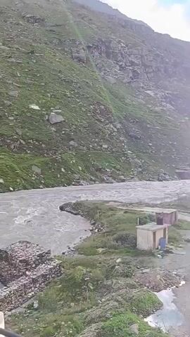 A wide shot shows a fast-flowing river in a mountainous region under a partly cloudy sky. On the left, a steep, rocky mountainside covered with green grass rises sharply. A dirt road runs parallel to the river, with several small, simple structures, possibly toilets or sheds, located near its edge. A few trucks are parked in the distance on the right side of the road. The sun is partially visible, shining brightly through the clouds and casting a lens flare onto the scene.