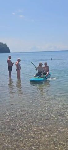 A man and a woman stand in shallow water near a pebbly beach. Two boys are on a paddleboard in slightly deeper water. One boy is paddling the board with a paddle, and the other boy is sitting behind him. The sky is clear and blue, and the water is calm and clear. In the distance, another person is swimming.