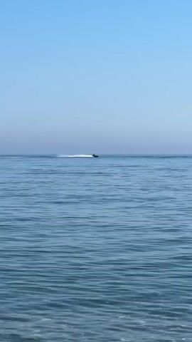 This media file shows a jet ski moving across the surface of the sea. The scene is set on a clear day with a bright blue sky. The water is a calm, deep blue, with small ripples on the surface. The jet ski is positioned in the middle of the frame, leaving a white spray of water in its wake. The bottom of the frame shows the clear, shallow water near the shore with pebbles visible on the seabed. The overall mood is peaceful and serene, suggesting a pleasant day at the beach. The location is identified as El Velerín, Spain. There is no visible text in the media file.