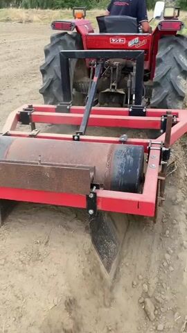 A red tractor is shown from a low angle from behind as it moves forward, digging into the dirt with an attachment. The attachment is red and metal with a large roller at the top and two V-shaped blades digging into the ground underneath the roller. The blades are pushing dirt outward, creating a trench. The tractor is in a dirt field with some vegetation visible in the background. A person is seated in the tractor's driver's seat.