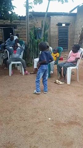 Here is a detailed description of the video:

The scene takes place in an outdoor courtyard, likely in a residential area. The ground is covered in fine, light-brown gravel or sand.

Several children are present. Some are seated on white and green plastic chairs arranged in a semi-circle. One child is standing in the center facing away from the camera. They are wearing a dark blue shirt and patterned blue jeans.

Buildings are visible in the background. They appear to be simple brick structures with corrugated metal roofing. There are trees and some large cactus-like plants also visible behind the children.

The general atmosphere is casual and informal, suggesting a neighborhood gathering or a play session among friends.