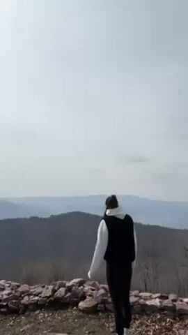 A man and a woman are standing on a stone wall with a view of rolling hills and mountains. The man is wearing a gray Nike sweatshirt and black pants. The woman is wearing a white hoodie, black vest, and black leggings. The man takes the woman's hand and pulls her in for a hug and a kiss. The sky is cloudy.