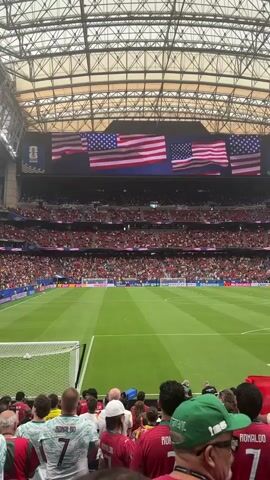 This media file captures a vibrant scene at a soccer stadium, likely in Houston, United States, given the prominent display of American flags on a large overhead screen. The stadium is filled with a diverse crowd of spectators, many of whom are wearing jerseys with the name "RONALDO" and the number "7" printed on the back, suggesting a connection to the famous soccer player Cristiano Ronaldo. The atmosphere appears to be one of excitement and anticipation, possibly for a match or a significant event.

The green soccer field is meticulously maintained, with white lines clearly demarcating the playing area and a soccer goal visible on the left side. The stadium's roof structure is visible, with a grid of metal beams and translucent panels that allow natural light to filter in, indicating it's daytime. The crowd is packed into the tiered seating, creating a sea of red and white in the foreground, with other colors interspersed throughout. The large screen above the field displays multiple waving American flags, adding a patriotic element to the event. Some spectators are holding up cell phones, likely to capture the moment, indicating the significance of the occasion. The overall impression is that of a large-scale sporting event with a passionate fanbase.