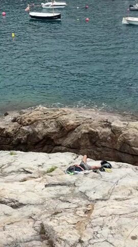 The video shows a person lying on a rocky outcrop near a body of water. The person is wearing shorts and appears to be resting or sleeping. The water is calm and there are several boats anchored in the distance. The overall scene is one of a peaceful, seaside environment.