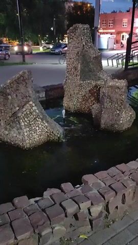 The video shows a stone fountain in a pond at dusk. A person on a bicycle is seen riding past the fountain in the background. Cars are parked and driving on a street behind the fountain, and a building with illuminated windows is visible across the street. The fountain itself is made of large, rough-hewn stones covered in smaller, light-colored pebbles. Water is spraying from the center of the fountain into the pond, which is filled with murky water and some green algae. A low brick wall borders the pond in the foreground. The lighting suggests it is either early morning or late evening.
