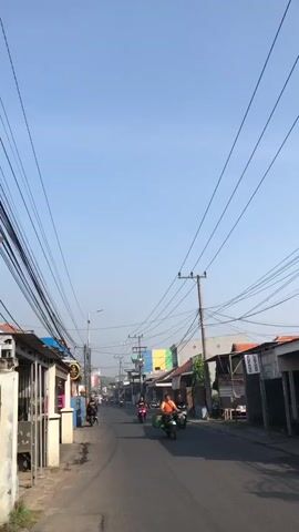 A clear, bright, daytime video displays a street scene with buildings lining both sides of a paved road. Overhead, a dense network of electrical wires crisscrosses the sky, supported by numerous utility poles. The road is populated by several motorbikes. One motorbike in the foreground is ridden by a man wearing an orange shirt and black pants, carrying a green box on the back. Another motorbike further down the road is ridden by a person in a red top. In the background, more motorbikes can be seen. The buildings appear to be small shops or residences. The sky is a uniform pale blue.