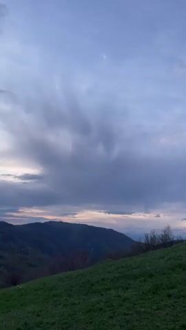 The video shows a green hillside with several white sword monuments and one concrete block monument. There are trees along the hillside. In the background, mountains are visible under a cloudy sky. Two people can be seen at the top of the hill, standing near the concrete block monument.