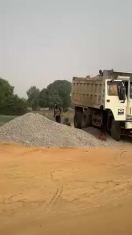 A long shot shows a white Hovo brand dump truck, apparently stuck in a large pile of gravel. A man is near the front of the truck, his body between the vehicle and the gravel pile. A second man is at the back of the gravel pile, throwing gravel with a shovel. The background is a sparse, arid landscape.
