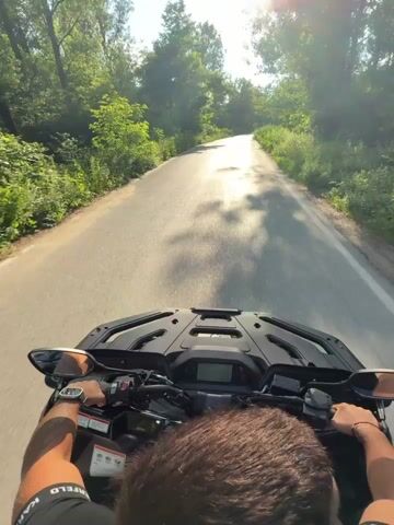 This is a first-person perspective video of someone riding an ATV on a paved road. The ATV is black and the rider's hands are visible on the handlebars, wearing a watch and a bracelet. The rider is wearing a black t-shirt with white text that appears to be "KARFELED".

The scene is a rural road surrounded by lush green trees and vegetation. The sun is shining brightly, indicating a daytime scene, likely late morning or early afternoon, with a clear blue sky. The road appears to be in moderate condition, with some cracks and unevenness visible. There are white lines marking the edges of the road and a white line down the center, though the center line is broken in some areas.

The road winds through a wooded area, and the sunlight creates bright highlights and shadows. In the distance, the road curves upwards and seems to lead towards a bridge or overpass. There are some signs visible on the side of the road, including what appear to be railway crossing warnings. The overall mood is one of outdoor recreation and enjoying a scenic drive. The location is identified as Boltata, Bulgaria.