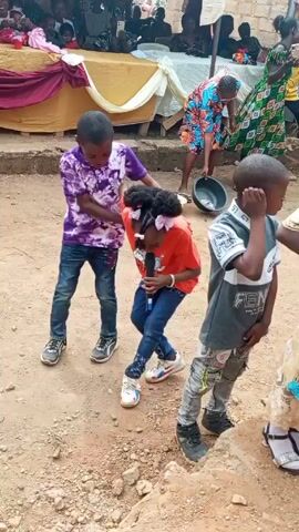 The video shows three children standing outside, with a crowd of people and a tent in the background. The child in the middle is holding a microphone and singing or speaking. The child to the left is touching the child with the microphone on the back. The child to the right is wearing a grey shirt with the words “FEN...” on it. The ground is dirt and gravel. The crowd in the background is watching and some people are holding up phones.