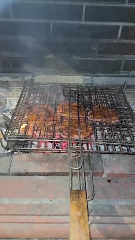 A grill is loaded with several pieces of seasoned meat, likely steaks or burgers, cooking over hot coals. The meat is dark brown and appears to be developing a crust. Smoke rises from the grill, indicating the cooking process. The grill is placed within a brick fireplace or outdoor grill structure. The camera pans slightly, showing the texture of the brickwork and the glowing embers beneath the meat. The overall scene depicts the preparation of food on a barbecue.