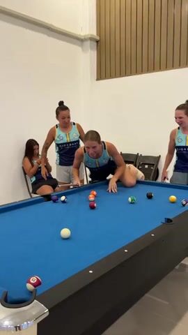 Four young women are playing pool in a room. The woman in the center is in a very low, almost contorted position, aiming to hit a red pool ball with her cue stick. She is wearing a dark blue and teal sleeveless athletic top and gray shorts. Two other women, also wearing similar athletic tops, stand behind her. One of them is looking at her phone. The woman to her right is looking at the player and smiling. A fourth woman is visible sitting on a chair in the background on the left. The pool table is covered in blue felt and has several balls scattered on it. The room has white walls and a slatted wooden ceiling.