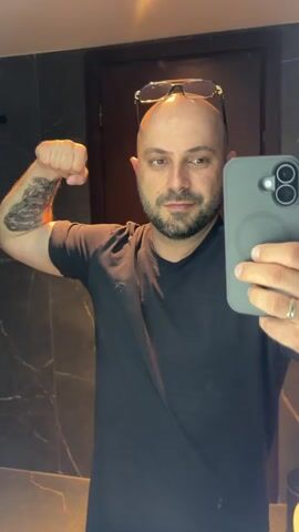 The video shows a bald man with a beard wearing a black t-shirt and sunglasses on top of his head. He is in a bathroom, reflected in a mirror. He is flexing his bicep, showcasing a tattoo on his arm. He holds a smartphone in his right hand, taking the selfie. A ring is visible on his left hand. The bathroom features dark, marbled tiles on the walls and a wooden door in the background. The lighting suggests it could be daytime or artificial lighting within an indoor space. The man's expression appears confident and proud. There is no visible text in the media.
