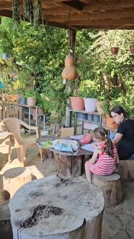The video shows a outdoor setting with lush greenery and potted plants. A woman and a young girl are sitting at a wooden table made of a tree stump, engaged in an activity involving papers and art supplies. The woman is wearing a black shirt, and the girl is in a striped dress. Several other wooden tree stump seats are visible around the area. Hanging dried herbs or plants adorn the overhead structure. The background features more foliage, suggesting a natural and serene environment. The camera pans around the outdoor seating area, showcasing the various potted plants and the natural surroundings.
