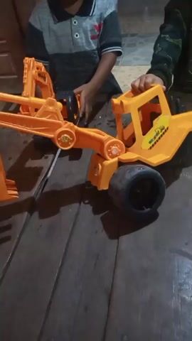Two young children are playing with toy construction vehicles on a wooden floor. In the foreground, a bright orange toy excavator is being manipulated by a child wearing a striped shirt. To the right, another child, partially visible and wearing a camouflage shirt, is interacting with a similar orange toy truck. The toys appear to be made of plastic. The lighting suggests an indoor setting, possibly a home. There are some small white particles scattered on the floor, possibly debris from the toys or the environment. The children are actively engaged with their toys, their hands and arms are visible as they move the vehicles.