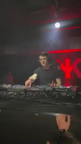 Here is a concise description of the video:

The video shows a DJ performing in a club. He's wearing a black t-shirt and headphones, manipulating the equipment in front of him. Other DJ equipment and various cables are visible on the table.  A partially visible logo, resembling "NYK", in red, is visible in the background. People are visible in front of the DJ booth, with hands raised. The room is dimly lit with some colored lights.
