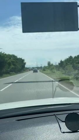 This media file is a first-person perspective video taken from inside a car driving down a two-lane road. The road is paved and has white lane markings. On either side of the road are dense green trees and vegetation, suggesting a rural or semi-rural setting. In the distance, there are utility poles and some low-lying buildings. The sky is blue with scattered white clouds, indicating it is daytime and likely fair weather.

The primary activity is driving. There are other cars visible on the road ahead, with one car in particular being the focus as it is directly in front of the filming vehicle. A blue road sign is visible on the right side of the road with text in Georgian. The visible text on the sign reads "სუფსა" and "ფოთი," which translates to "Supsa" and "Poti." These are place names in Georgia, providing a specific location context. The overall mood is neutral, reflecting a typical car journey. The time of day appears to be midday or early afternoon due to the bright sunlight and clear sky.