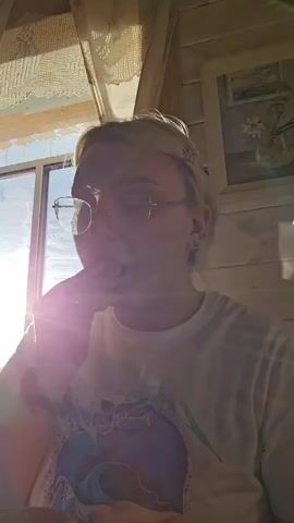 A young person with blonde hair, wearing glasses and a white t-shirt, is shown from the chest up, sitting indoors near a window. The sun is shining brightly through the window, creating a lens flare effect and casting a warm glow on the scene. The person is holding a mug with both hands and appears to be drinking from it. The background suggests a rustic interior with wooden walls and a lace curtain. A framed picture of flowers is visible on the wall. The person's expression is neutral, and the overall mood is relaxed and serene, possibly suggesting a morning or afternoon setting due to the bright sunlight. No visible text is present in the media.