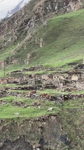 The video shows a hillside with the ruins of stone buildings. Green grass covers the ground. Some of the stone structures are in better condition than others, with one appearing partially renovated with a red roof. A utility pole stands nearby. The hillside slopes upwards towards a mountain in the background, part of which is covered in snow.