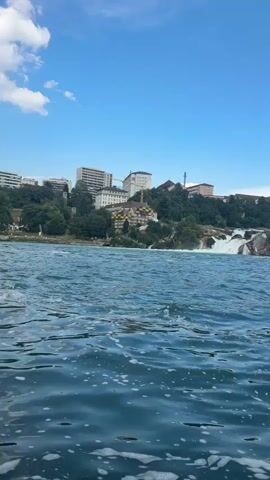 This is a video clip of the Rhine Falls in Neuhausen am Rheinfall, Switzerland. The camera is positioned low on the water, looking towards the falls. The water in the foreground is choppy with white foam, suggesting movement or waves.

In the background, the Rhine Falls are visible, a wide cascade of white water plunging over rocks. The falls are surrounded by lush green trees and rocky cliffs. Above the falls and trees, a town or city is built on the hillside, with several multi-story buildings, including what appears to be a large hotel or administrative building with a prominent "Casino" sign. Further up the hill, a picturesque castle or historic building with turrets stands out, nestled amongst the trees. The sky is a clear, bright blue with a few wispy white clouds, indicating fair weather. The sun is likely high in the sky, suggesting it is midday. The overall scene evokes a sense of natural beauty and human settlement coexisting. There are no people clearly visible in the frame.