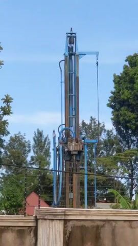 The video shows a blue water well drilling rig in operation, surrounded by trees and a fence. The rig has a tall, vertical frame with a drilling head and various hoses and cables. The drilling head moves up and down, driving a drill bit into the ground. The video is shot from a distance, and there are no people visible in the frame. The background includes a blue sky and some buildings. The overall impression is of an industrial process taking place in a semi-rural environment.