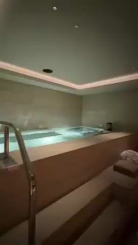 The video shows a modern spa room featuring a square jacuzzi, a stepped wooden bench, and a sauna. The jacuzzi is filled with water, and a towel is placed on the bench. The sauna, visible through a glass wall, has multiple levels of wooden seating illuminated by warm, indirect lighting. The room has soft, ambient lighting from above, highlighting the clean lines and neutral tones of the space.
