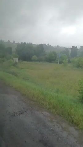 The video shows a drive through a rural landscape under an overcast sky. The road is wet, suggesting recent rain. Several road signs are visible: a speed bump sign and a pedestrian crossing sign. The surrounding area is characterized by green fields and scattered trees, with some small buildings and houses visible in the distance. The overall atmosphere is subdued, likely due to the cloudy weather. No people are clearly visible, and there are no apparent interactions or significant events occurring.