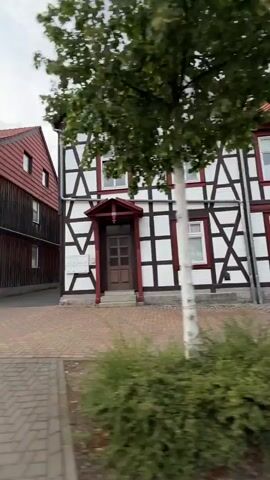 The video shows a narrow cobblestone alleyway between two traditional German half-timbered houses. The house on the left is painted light beige with dark brown wooden beams. It has a red tiled roof and several windows. The house on the right features a more ornate half-timbered facade with white plaster and dark brown beams, and a red trim around some windows and the entrance. A car is partially visible in front of the left house. The alleyway opens into a small courtyard with greenery and another building in the background. The sky is overcast. The camera pans to the right, revealing more of the greenery and foliage bordering the alley and houses.