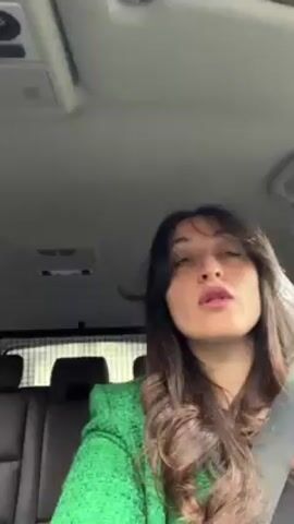 A woman with long brown hair and a green top is in the driver's seat of a car. She is wearing a seatbelt. She glances up and to the side. The car's interior is visible, including the ceiling, seats, and a safety screen behind the driver.