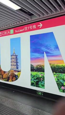 The video captures a bustling scene in a modern subway station in Gongshu District, China. The primary subjects are a diverse group of people walking along a concourse. They appear to be commuters or travelers, with varying ages and styles of dress. Some individuals are carrying bags, suggesting they are either shopping or on their journey. The mood seems neutral and everyday, typical of a public transit hub.

The setting is an underground subway station, evident from the tiled floor, overhead lighting panels, and the metallic railings. A prominent feature is a large, brightly lit digital advertisement spanning a significant portion of the wall. This advertisement displays large, stylized letters that form words, interspersed with scenic images of natural landscapes, traditional Chinese architecture, and vibrant skies. The imagery suggests a focus on local attractions or a promotion of the area's beauty.

Visible text on the advertisement includes: "Fengqi Metro Shops," "To Line1 往1号线," and also the letters "CBA" which might refer to different sections or services within the station. The overall impression is one of a well-maintained and modern urban environment, where public transportation infrastructure is integrated with visual displays intended to inform and perhaps entertain passengers. The time of day is not explicitly stated but the lighting suggests it's likely daytime, or at least during operating hours of the subway. There are no obvious indications of weather conditions as the scene is entirely indoors.