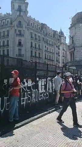 The video shows a street in a city with people standing near a black metal fence. The fence has graffiti on it that says "NO A LAS MINERAS." There are buildings visible behind the fence, including one that has "HOSTEL" written on it. There are some people holding cameras.  A man in a red hat stands facing the fence, while other people stand in groups and walk along the sidewalk.  The sky is clear and the sun is shining.